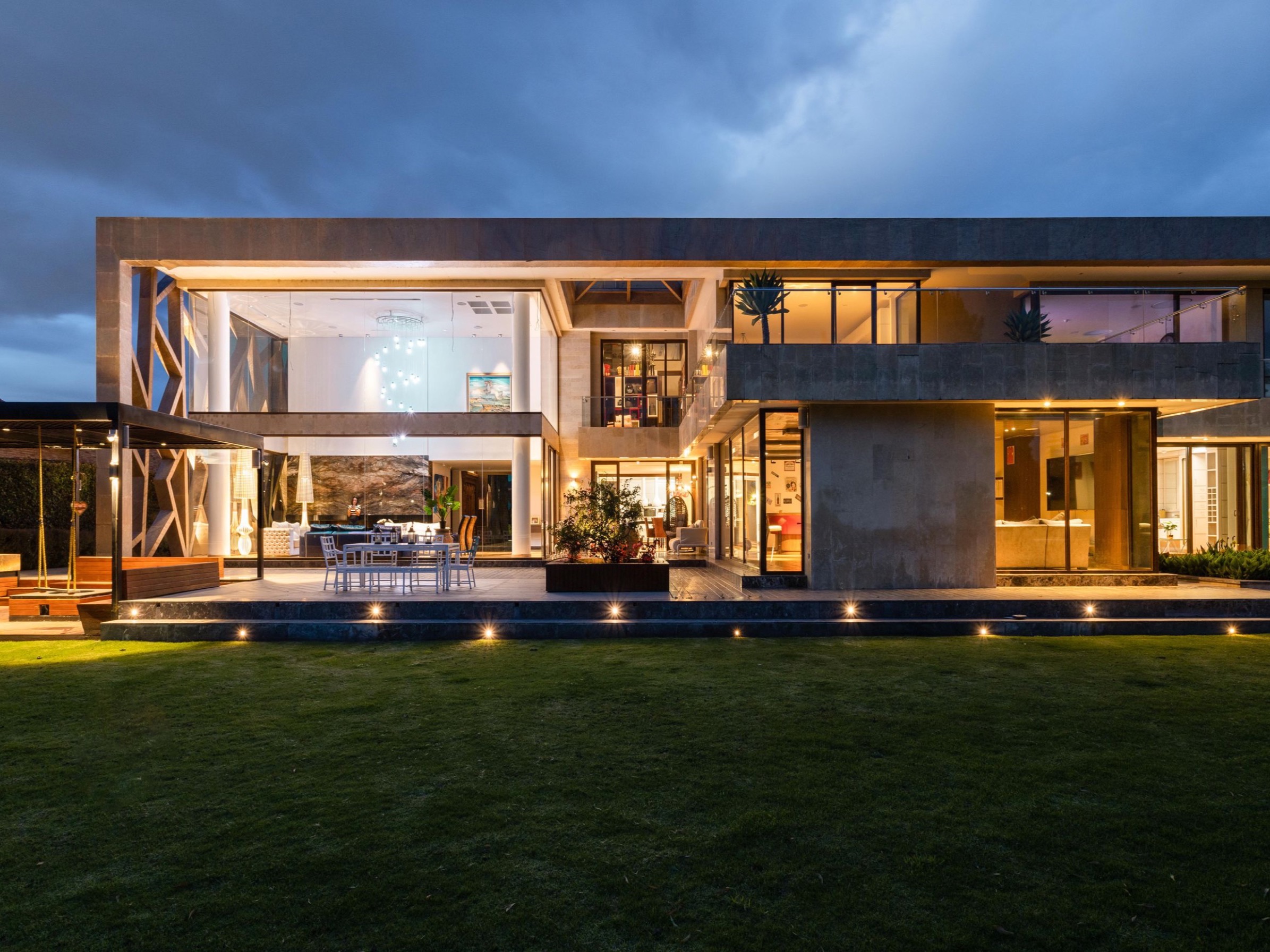 A low concrete and glass house at dusk, its whole ground floor glazed and lit warm, lawn running up to the terrace
