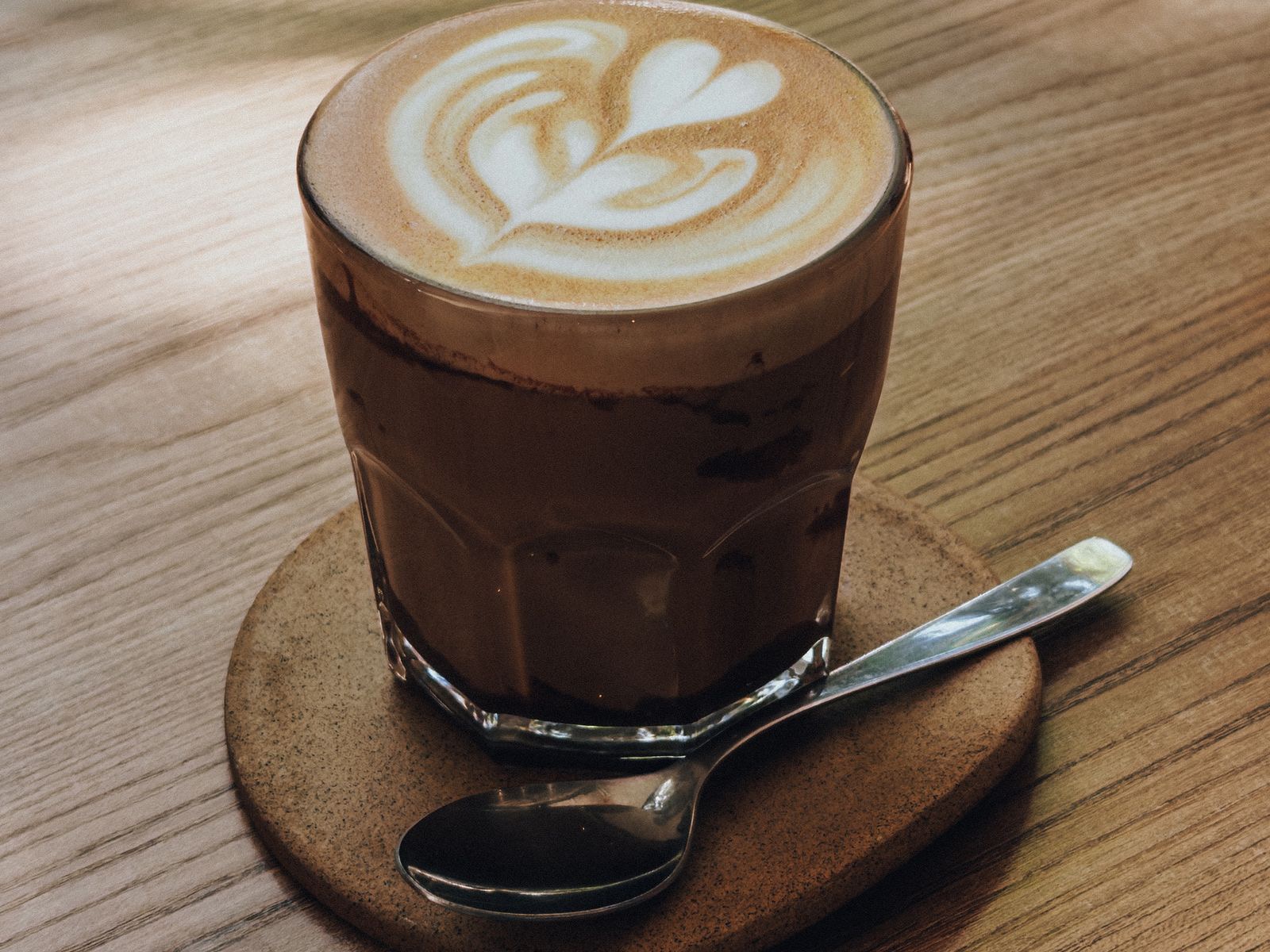 A flat white in a glass with latte art on top, on a wooden coaster